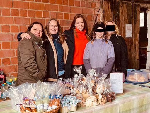 Fünf Personen stehen an einem Tisch mit Backwaren und Kunsthandwerk, umgeben von einer rustikalen Kulisse.